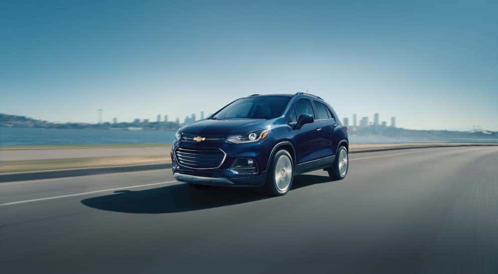 Dark blue 2018 Chevrolet Trax driving down a paved road past a body of water and a city skyline