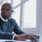 Man in a blue sweater over a white button-up sitting at a white desk using a white laptop