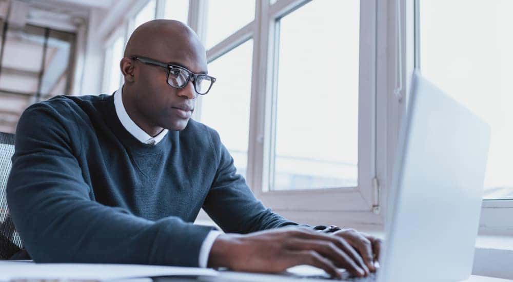 Man in a blue sweater over a white button-up sitting at a white desk using a white laptop