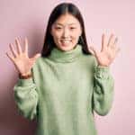 Woman with black hair in a light green sweater holding ten fingers up in front of a pink wall