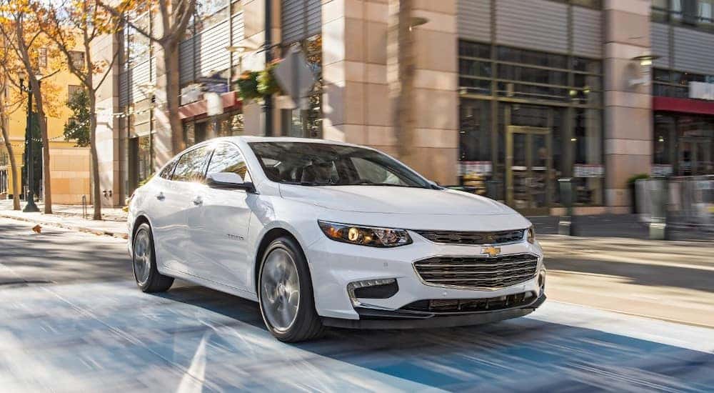 White 2018 Chevrolet Malibu sedan driving down a paved road past a brick building