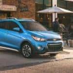 Light blue 2018 Chevrolet Spark parked in front of a brick building and next to tables with umbrellas on them