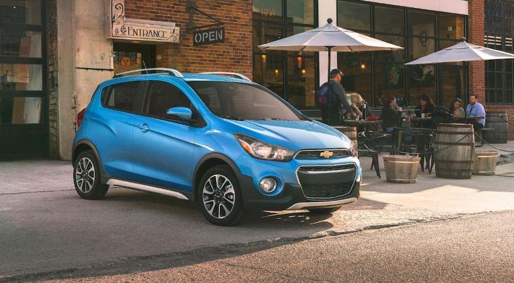 Light blue 2018 Chevrolet Spark parked in front of a brick building and next to tables with umbrellas on them