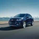 Dark colored 2018 Chevrolet Trax SUV driving on a paved road past a body of water and tall city buildings