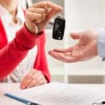 Woman in an orange cardigan handing over a car key to a man in a suit with a contract and clipboard on a desk below their hands