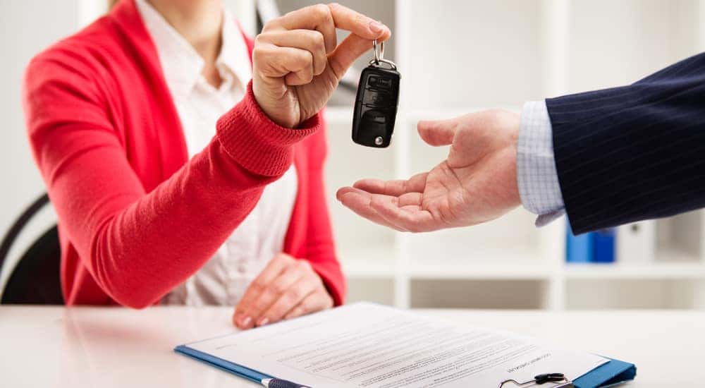 Woman in an orange cardigan handing over a car key to a man in a suit with a contract and clipboard on a desk below their hands
