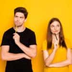 Man in a black shirt and woman in a yellow shirt standing in front of a yellow wall with pencils to their chins, thinking