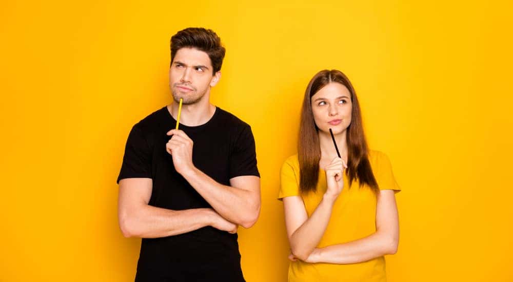 Man in a black shirt and woman in a yellow shirt standing in front of a yellow wall with pencils to their chins, thinking
