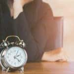 Woman in a black shirt with her elbows on a table and hand to her chin with a clock in front of her
