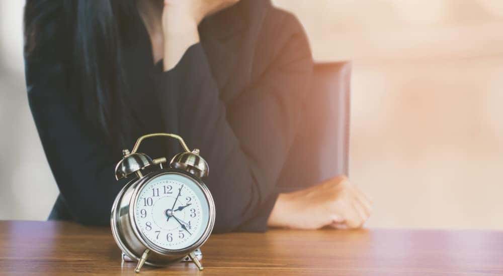 Woman in a black shirt with her elbows on a table and hand to her chin with a clock in front of her
