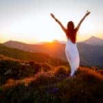Woman in a white dress standing on a mountain with her hands raised to the sunset and distant mountains