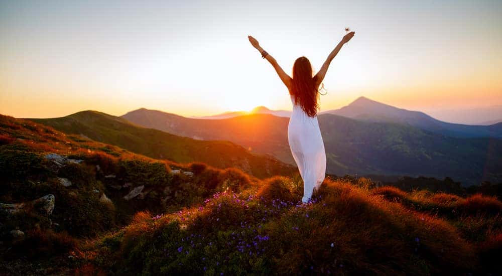 Woman in a white dress standing on a mountain with her hands raised to the sunset and distant mountains