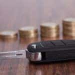 Close-up of a car key on a brown table in front of consecutively taller stacks of coins