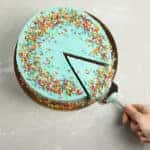Hand using a spatula to take out a single slice of a blue cake with sprinkles all over it