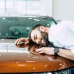 Man in a button-up shirt with his head, arms, and upper body on the hood of an orange car, dreaming