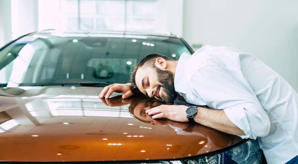 Man in a button-up shirt with his head, arms, and upper body on the hood of an orange car, dreaming