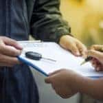 Man holding out a clipboard and car key while woman signs a paper with a black and gold pen