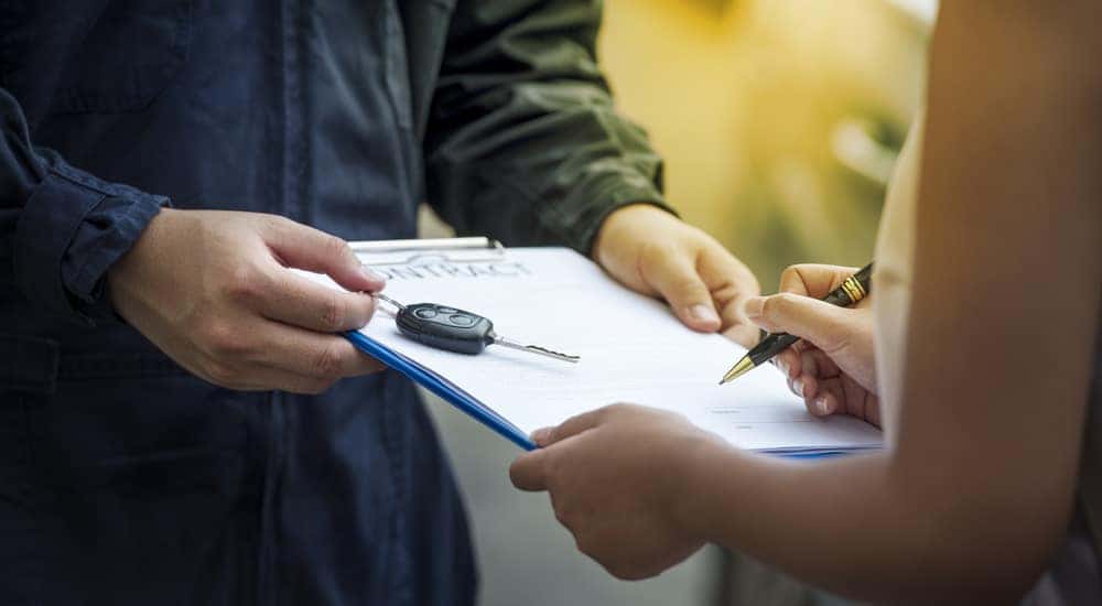 Man holding out a clipboard and car key while woman signs a paper with a black and gold pen