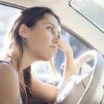 Woman with brown hair in a tank top leaning against the wheel in her car, thinking deeply