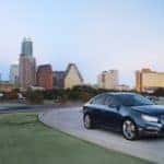2015 Chevrolet Cruze sedan driving down a paved road with green grass on either side and tall city buildings in the background