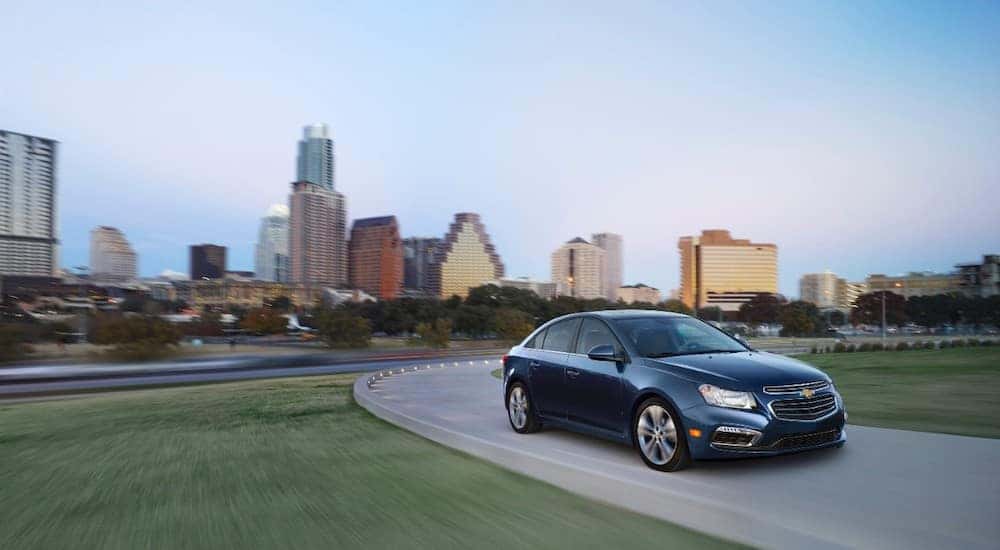 2015 Chevrolet Cruze sedan driving down a paved road with green grass on either side and tall city buildings in the background