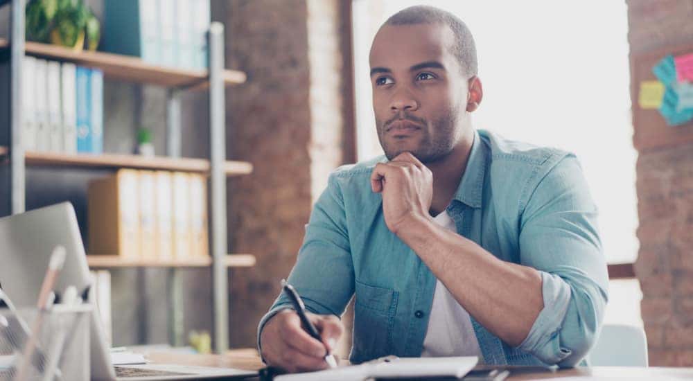 Man in a blue shirt sitting with his hand on his chin at a desk with a laptop and notebook