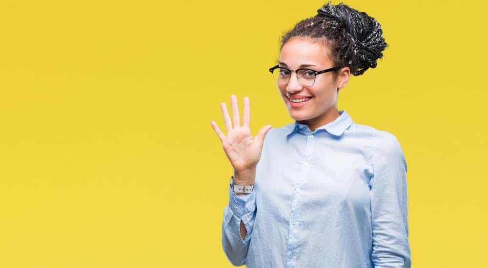 Woman in a blue button-up holding five fingers up in front of a yellow wall
