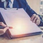 Man in a suit holding up a contract on a clipboard on a desk with car keys sitting on it