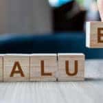 Hand placing an "E" letter block on a grey table next to other blocks to spell out the word "VALUE"