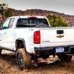 Back view of a white 2018 Chevrolet Colorado with mud on the tires on a dirt path in front of mountains