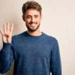 Man in a blue sweater with four fingers up, smiling in front of a white wall