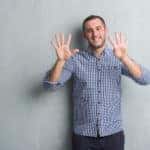 Man in a blue button up shirt holding up nine fingers in front of a grey wall