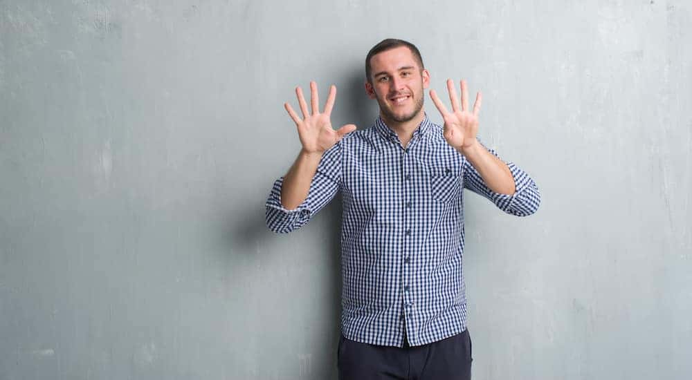 Man in a blue button up shirt holding up nine fingers in front of a grey wall
