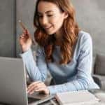 Girl in a light blue shirt holding a pencil, using a laptop, and sitting at a table with open books on it