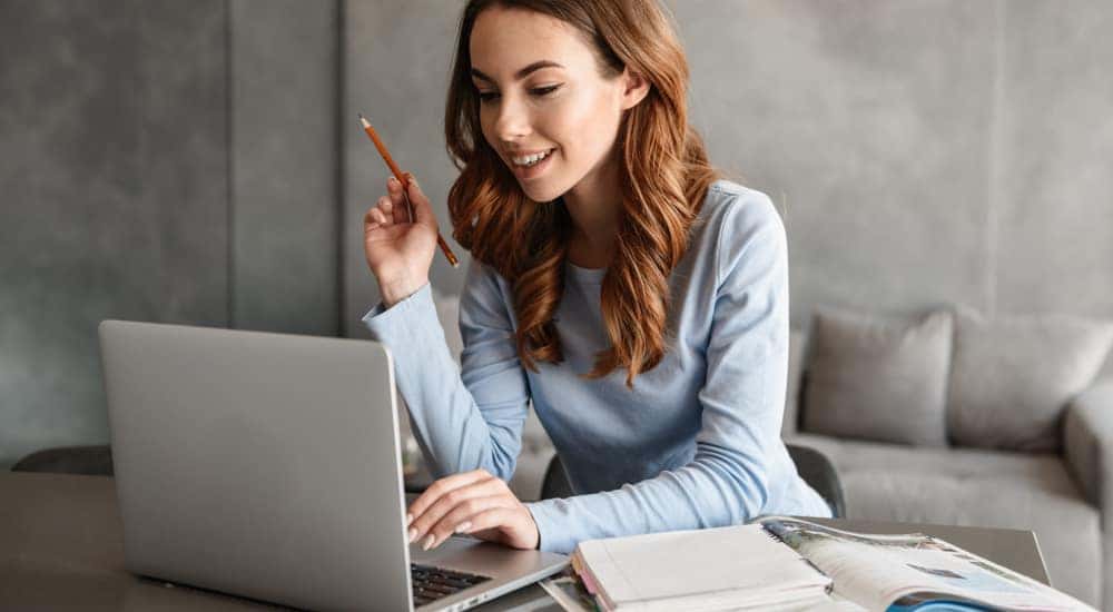 Girl in a light blue shirt holding a pencil, using a laptop, and sitting at a table with open books on it