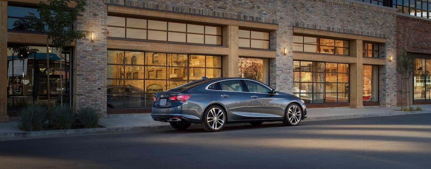 A dark grey 2020 Chevy Malibu, a popular car at Chevy dealerships near Clifton Park, NY, is parked in front of a brick building at night.