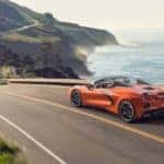 Back to front aerial view of an orange 2020 Chevrolet Corvette driving down a road toward a mountain and next to a body of water