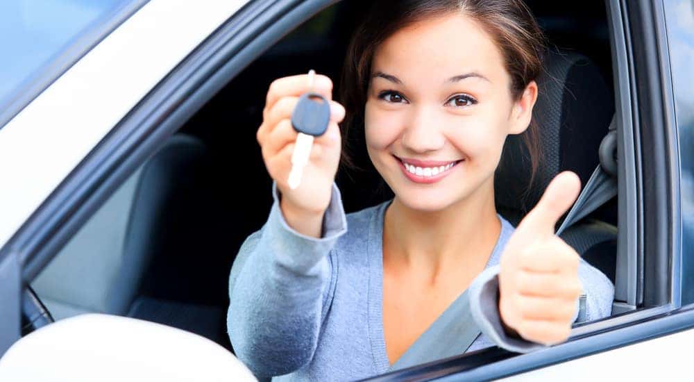Teen girl sitting in the driver's seat of a white vehicle with the window open holding up a car key and a thumbs up