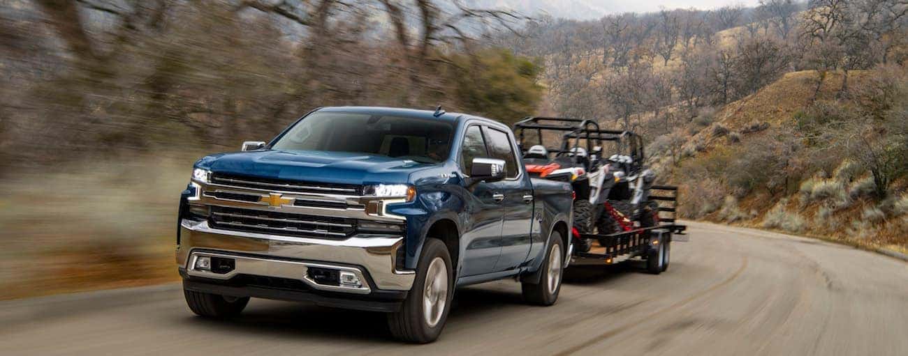 A blue 2020 Chevy Silverado 1500 is towing a trailer loaded with two side by sides on a dirt road near Albany, NY.