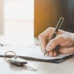 Hand holding a pen and signing a car lease with car keys next to it