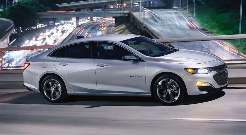 A silver 2019 Chevrolet Malibu, which is a popular used car at car dealerships near me, is driving over a busy highway at night.