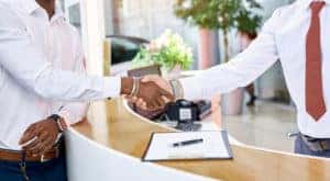 Two men in button-up shirts shaking hands over a counter and clipboard in a dealership showroom