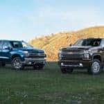 A blue 2019 Chevy Silverado 1500 is next to a dark grey 2020 2500HD in front of a hill.