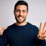 Man with dark hair and facial hair in a blue sweater holding eight fingers up in front of a grey wall