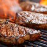 Three steaks sitting on a grill with flames coming through