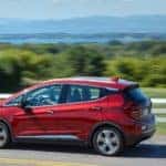 Red 2020 Chevrolet Bolt EV driving down a paved street next to a white fence, green grass and trees, and tall mountains