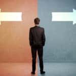 Man in a black suit standing in front of a half orange, half blue wall with arrows facing in opposite directions