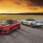 One red 2021 Camaro and one silver 2021 Camaro parked on pavement with flames behind them