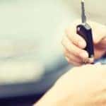 A hand is giving a car key to another hand with a car in the background.