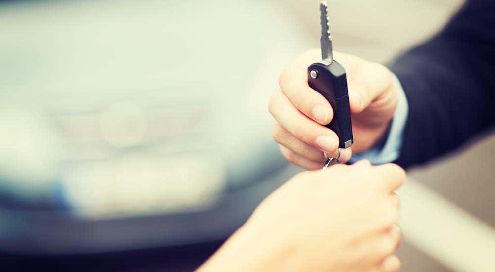 A hand is giving a car key to another hand with a car in the background.